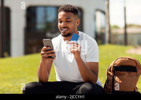 Positive black man holding credit card and smartphone sitting on bench in the city park, making purchase in online store Stock Photo