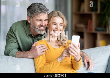 Portrait Of Smiling Middle Aged Couple Resting With Smartphone On Couch Stock Photo
