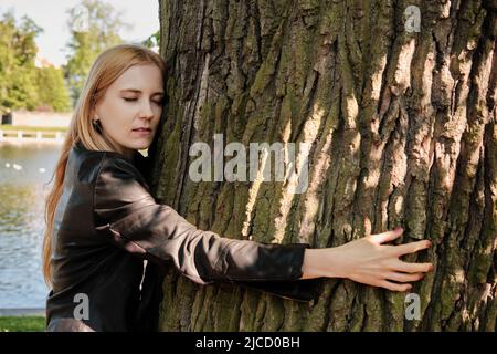 Pretty woman with long blond hair, wearing a leather jacket, hugging a giant oak tree trunk. Ecology concept. Stock Photo