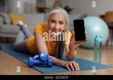 Fitness mobile app. Happy senior woman showing smartphone with blank screen, exercising at home, mockup Stock Photo