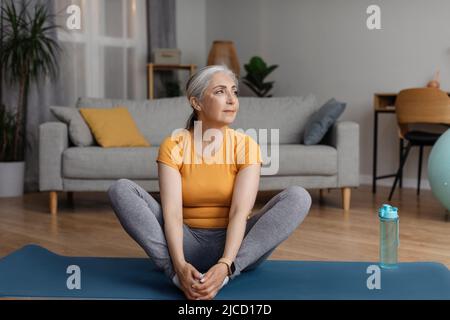 Aged woman sitting in butterfly yoga pose on sports mat at home, doing flexibility exercises, keeping fit and healthy Stock Photo