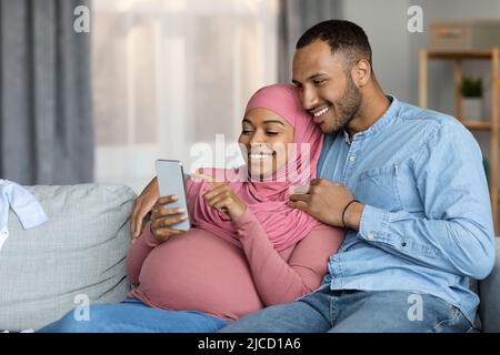 Happy Pregnant Black Islamic Couple Using Smartphone While Relaxing Together At Home Stock Photo