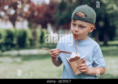 Child eating on the go pan-fried noodles chopsticks. Dirty soy sauce stains on a white t-shirt. Takeaway food. outdoors Stock Photo