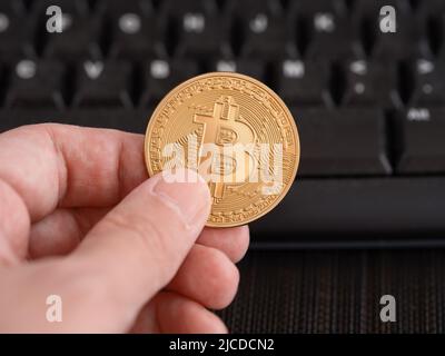 A man holding a golden bitcoin coin in his hand against a computer keyboard. Close up. Stock Photo