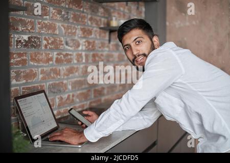 young man using a laptop and a smartphone standing in the kitchen. Stock Photo
