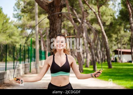Happy sportive woman wearing sports bra standing on city park, outdoors skipping rope jumping exercises. Healthy lifestyle, outdoor sports concept. Stock Photo