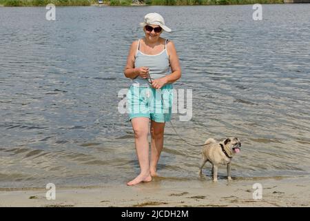 Smiling aged woman with pug on a leash are standing in shallow of river water on riverbank. Stock Photo