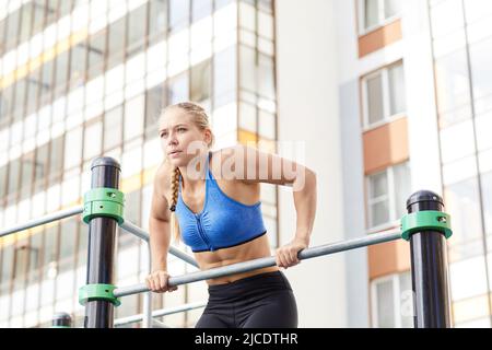 Motivated young woman in sportswear breathing out while doing chin-up exercise at workout area by house Stock Photo