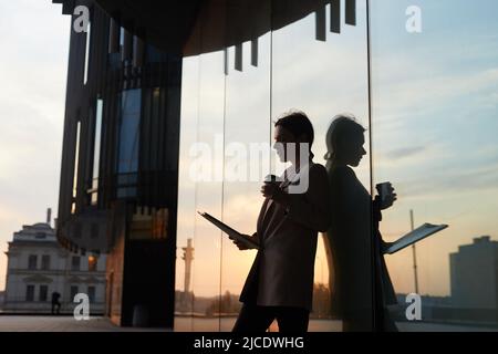 Busy contemporary lady in jacket standing by glassy building and drinking coffee while viewing papers outdoors, reflection effect Stock Photo