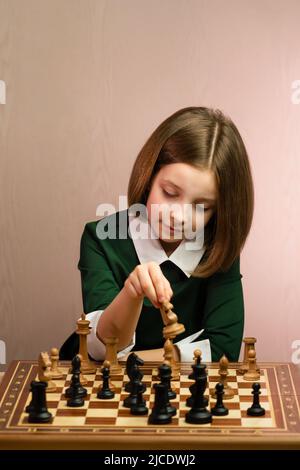 Satisfied Young Child Chess Player Makes Move with Knight in Chess Game. School Girl Sitting at Chessboard in Green Dress. Chess Opening with White Stock Photo