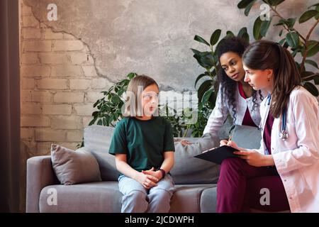 Doctor Pediatrician and Non Caucasian Skinned Woman Intern Getting Medical History from Child Girl in Medical Office. Female Patient Visits Pediatric Stock Photo