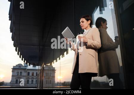 Below view of pensive attractive middle-aged lady in elegant jacket leaning on building wall and watching video presentation on tablet Stock Photo