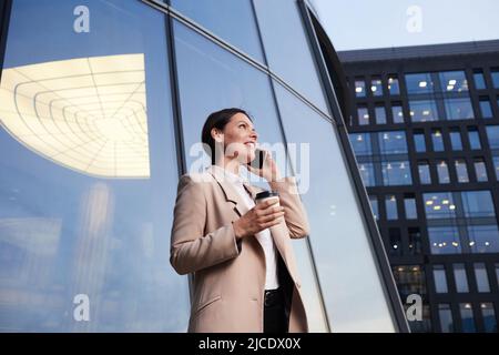 Positive successful business lady in long jacket standing by modern building and calling on phone while drinking coffee Stock Photo