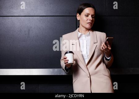 Thoughtful elegant middle-aged business lady in fashionable jacket leaning on railing in dark lobby and using smartphone while drinking coffee Stock Photo