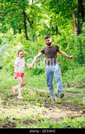 Portrait of happy family walking in park. Little cheerful daughter holding hand of middle-aged smiling bearded father. Stock Photo