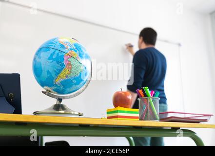Detail of a school classroom. Close-up of the teacher's desk with objects on it and in the background, out of focus, the teacher. Stock Photo