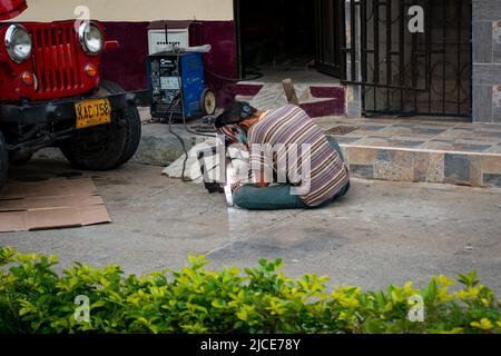 Cisneros, Antioquia, Colombia - February 20, 2022: Young Man Sitting on the Ground on the Street Welding a Piece of Iron next to a Car Stock Photo