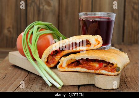 Two pieces of pizza calzone with glass of red wine, fresh scallion and tomato on wooden board horizontal Stock Photo
