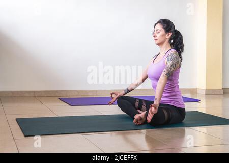 Seated adult latin woman with tattoos meditating and practicing yoga in pose padmasana Stock Photo
