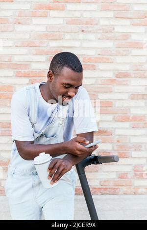 Content young African American male millennial in stylish outfit with dark hair and beard. He is leaning on kick scooter and smiling while messaging on smartphone standing on street with takeaway coffee Stock Photo