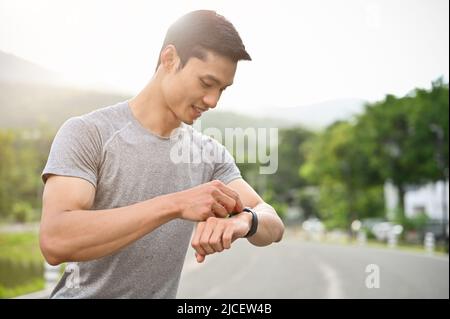 Handsome asian young athletic male in sportswear taking a break after running, checking his pulse and heart rate on a smartwatch. Sport and technology Stock Photo