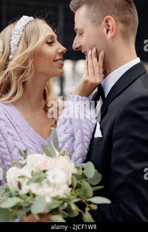 Beautiful young wedding couple in elegant outfits walk around city, embrace and kiss on street background. Portrait closeup of happy bride with Stock Photo