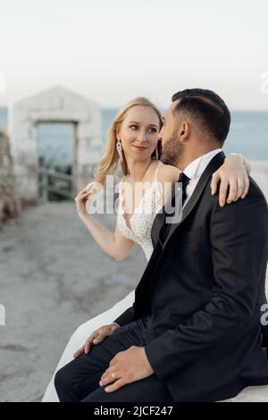 Portrait of young blonde bride in white dress looking at brunet groom in suit, embracing and sitting near stone arch on sea coast in Italy side view Stock Photo