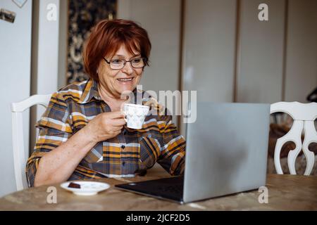 Smiling senior woman in glasses, using laptop at home. Drinking tea or coffee. Working at home, computer technology.  Stock Photo
