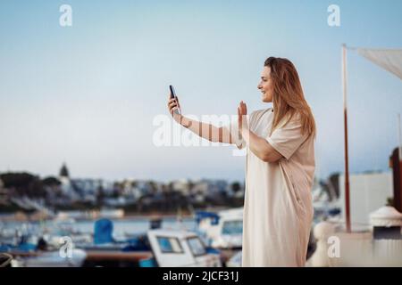 Glad and smiling blond long-haired pregnant woman looking at screen mobile phone, saying hi. Side view. Copy space Stock Photo