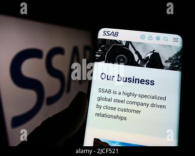 Person holding cellphone with webpage of Swedish steel company SSAB AB on screen in front of business logo. Focus on center of phone display. Stock Photo