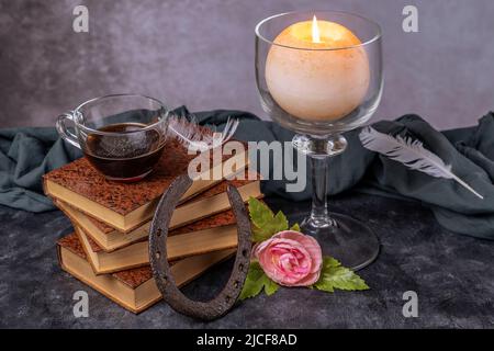A stack of old books with a glass cup with coffee on it, a horseshoe, a rose and a lighted candle Stock Photo