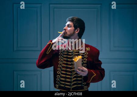 Comic portrait of young man in image of medieval hussar smoking fries isolated on dark blue background. Retro style, comparison of eras concept. Stock Photo