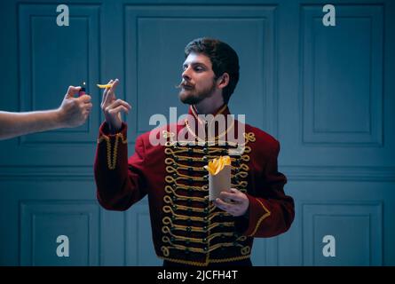 Comic portrait of young man in image of medieval hussar smoking fries isolated on dark blue background. Retro style, comparison of eras concept. Stock Photo