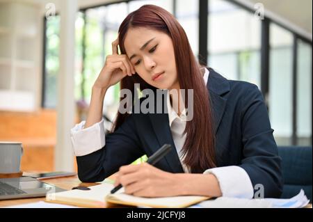 Stressed and bored asian female business employee worker feeling no passion with her work, sits at office desk and writing something on notebook. Upse Stock Photo