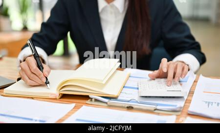 A businesswoman or financial assistant working at her office desk, analysing financial data, taking notes notebook, using calculator. cropped image Stock Photo