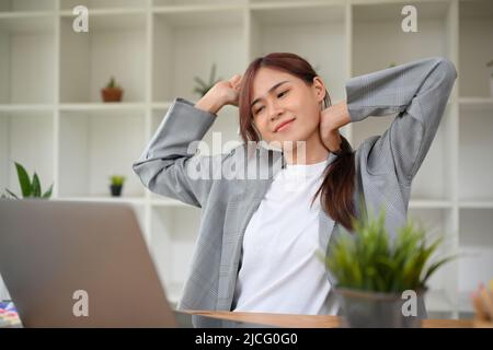 Beautiful and cheerful asian young businesswoman relaxing and stretching her arms and neck after work at her office desk. Stock Photo