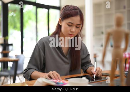 Attractive young asian female graphic designer sits at her office desk, designing her creative project, using graphic tablet. Stock Photo