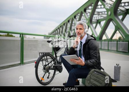 Businessman with bike sitting on bridge, using tablet and listening to music. Commuting and alternative transport concept Stock Photo
