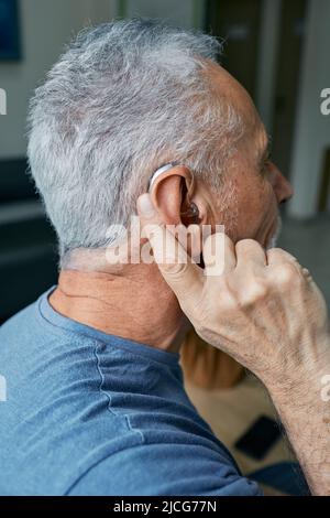 Senior man tunes his hearing aid behind the ear by pressing his finger on setting button. Modern hearing aid, hearing solutions Stock Photo