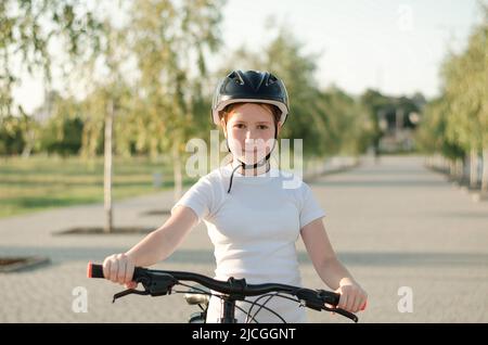 Teenage girl in a helmet on a bicycle. Red-haired girl in a white T-shirt on a bike ride in the park. Stock Photo