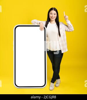 Asian woman pointing finger one as attention leaned back on giant, huge smartphone with blank white screen, wearing casual isolated on yellow background. Free space mock up. Stock Photo