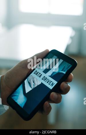 a man, wearing a white shirt and a gray suit jacket, holds his smartphone, that reads the text data driven in its screen, in his hand Stock Photo