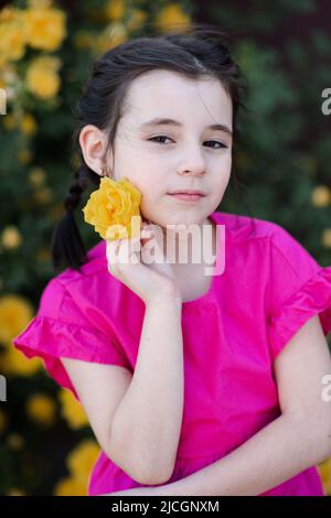 Stylish little kid girl 6-7 year old smelling yellow rose flower over blooming bushes in garden outdoor close up. Looking at camera. Springtime. Cute Stock Photo