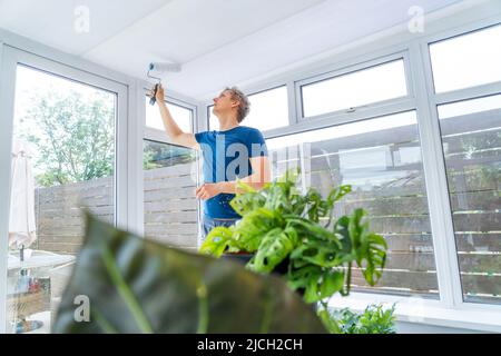 Young man painting the plastic ceiling of the conservatory in white color by the roller. DIY, Do it yourself concept. House improvement. Home renovati Stock Photo