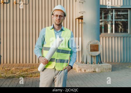 Foreman supervisor in suit and helmet on head, holding folder with documents at industrial factory. Chimney pipe of mini thermal power plant in backgr Stock Photo