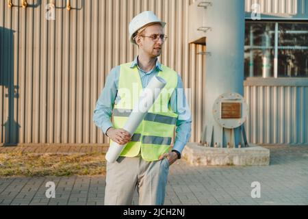 Foreman supervisor in suit and helmet on head, holding folder with documents at industrial factory. Chimney pipe of mini thermal power plant in backgr Stock Photo