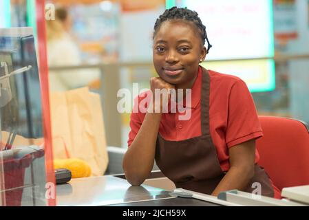 Young successful saleswoman in uniform sitting by cashier counter in supermarket and looking at camera while keeping hand by chin Stock Photo