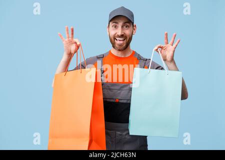 Smiling male courier in uniform holding shopping bags on blue studio background Stock Photo
