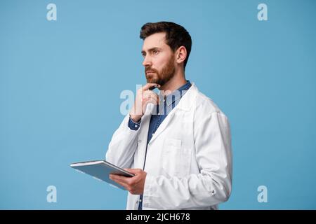 Focused man doctor in white coat stands with clipboard in hands and looks away Stock Photo