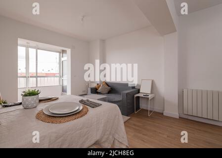 Living room and dining room with gray sofa, table with beige linen tablecloth and light wood flooring Stock Photo